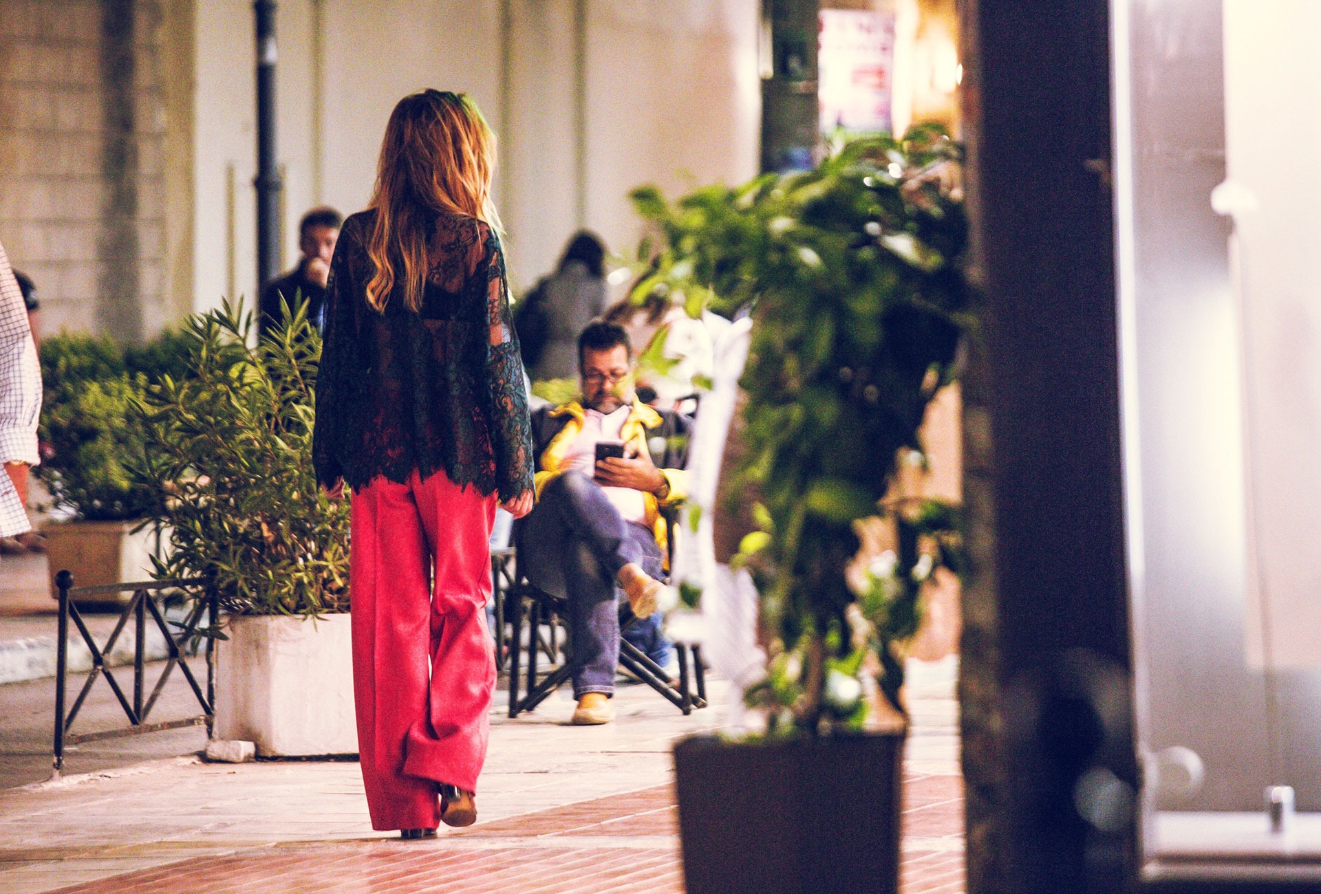 Kobieta w czerwonych spodniach i koronkowej bluzce idzie po chodniku, w tle siedzący mężczyzna z telefonem i rośliny w donicach