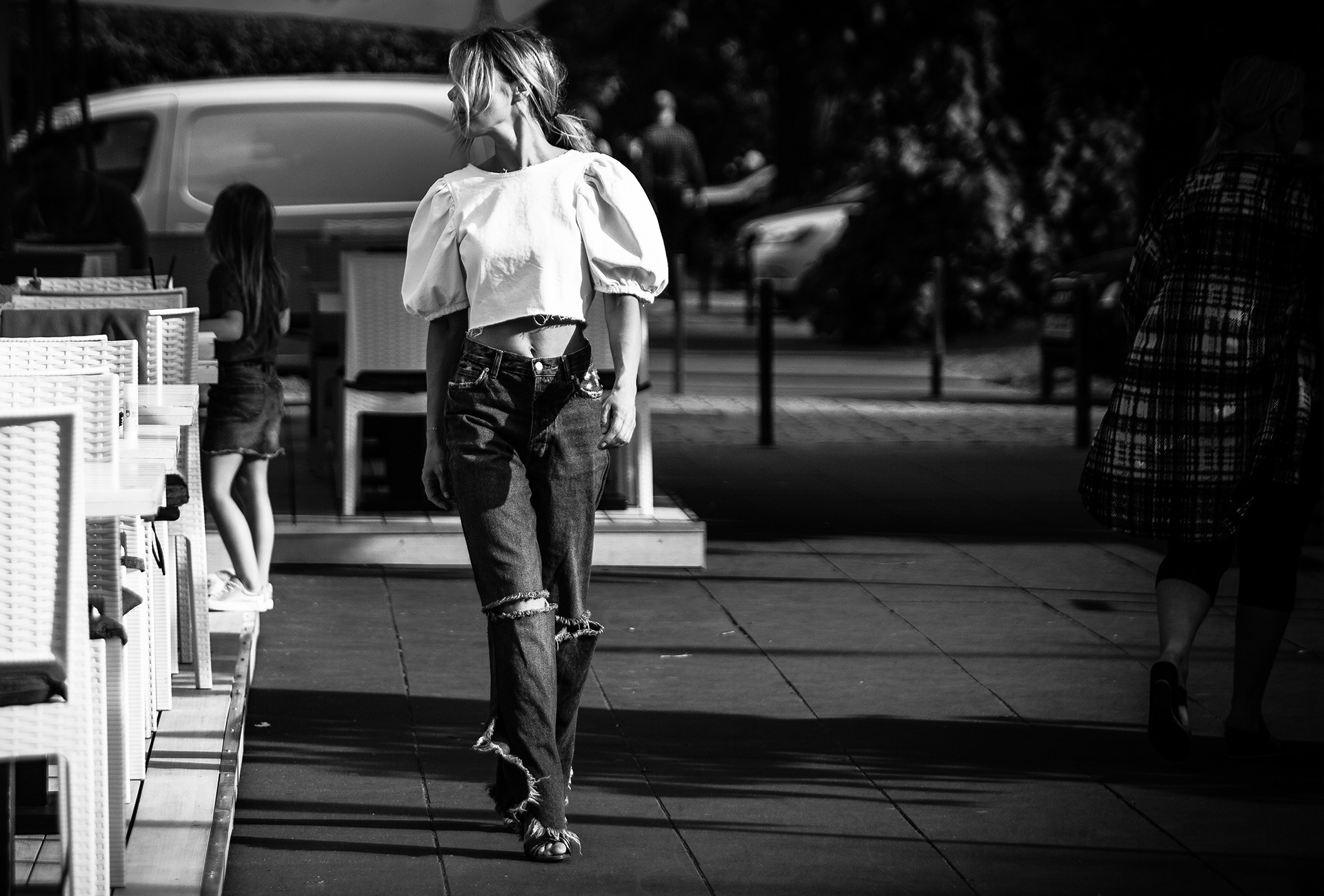 Zdjęcie czarno-białe przedstawia kobietę idącą chodnikiem w stylizacji z bluzką z bufiastymi rękawami i podartymi dżinsami, z widocznym fragmentem brzucha, odwróconą w lewo, z włosami spiętymi...