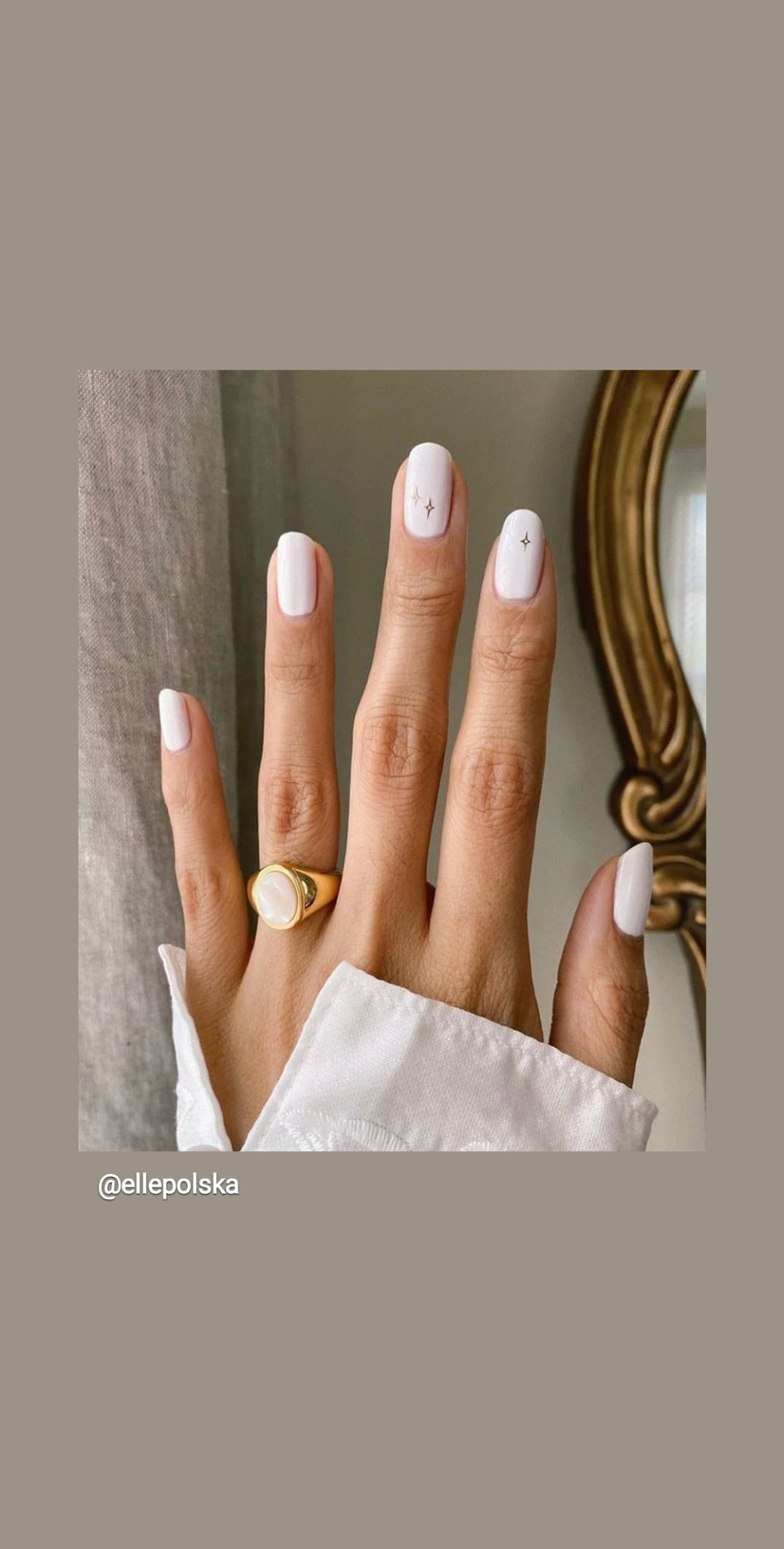 Dłoń z białym manicurem z subtelnymi złotymi zdobieniami w kształcie gwiazdek, ozdobiona złotym pierścieniem z perłowym oczkiem, na tle beżowej zasłony i fragmentu złotej ramy lustra.