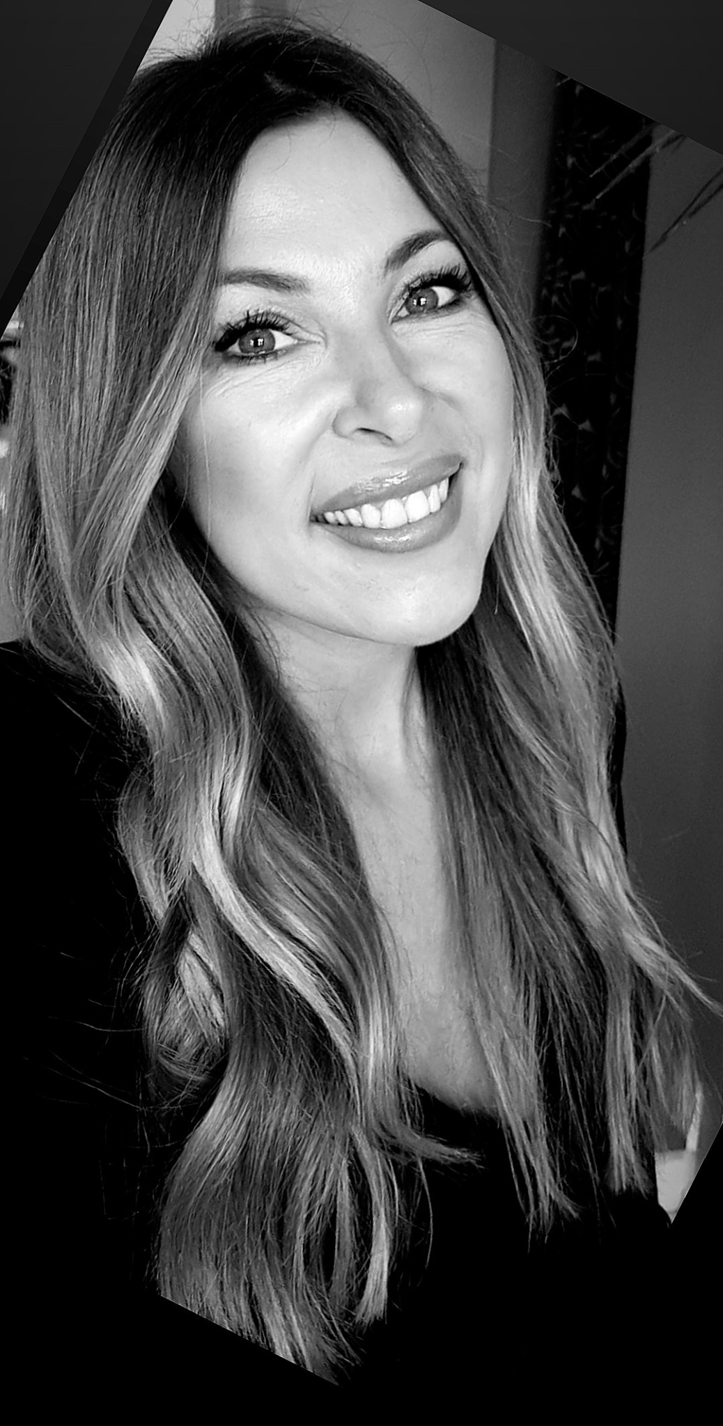 Portret uśmiechniętej kobiety z długimi, falowanymi włosami w odcieniach blondu, z delikatnym makijażem oczu i błyszczącymi ustami, w czarno-białej tonacji.