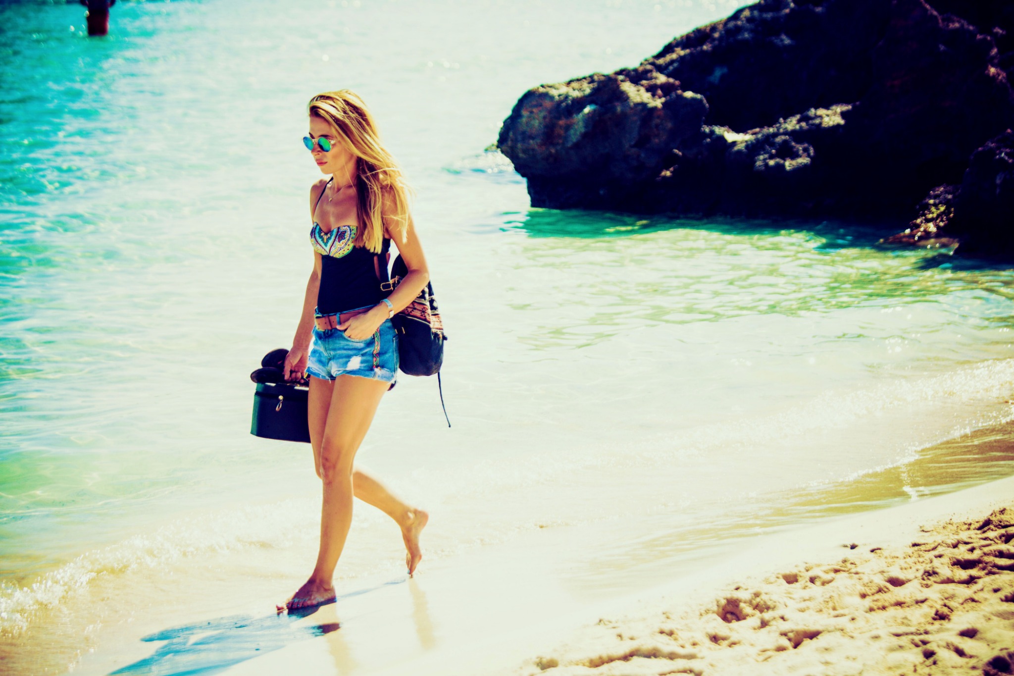 Kobieta w letnim stroju i okularach przeciwsłonecznych idzie brzegiem morza, niosąc czarną kosmetyczkę w ręku.