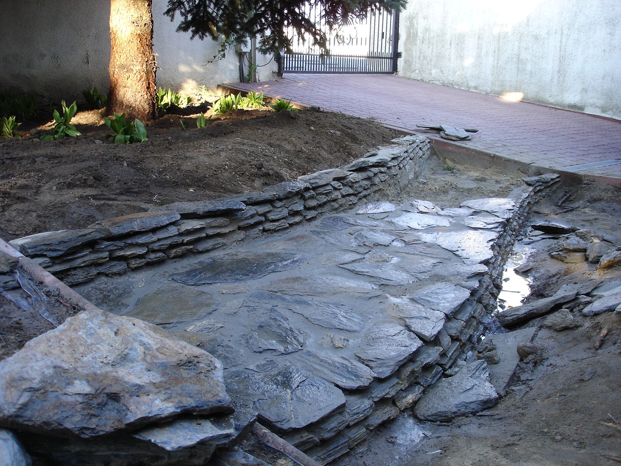 Budowa kamiennego koryta z nieregularnych płyt w ogrodzie, widoczne fragmenty murku z łupka, ziemia, częściowo ułożona kostka brukowa i brama w tle.