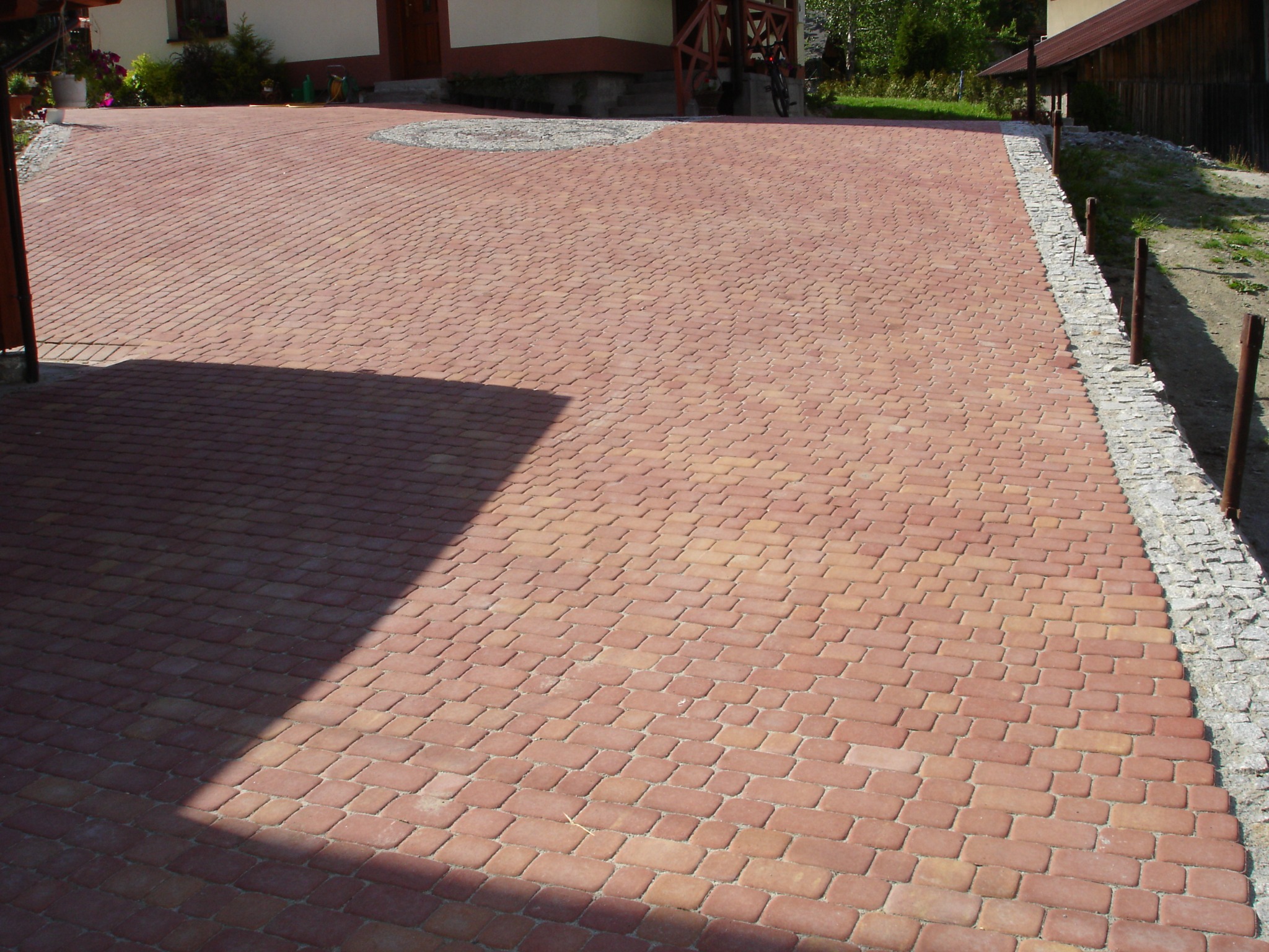 Nowa, czerwona nawierzchnia z kostki brukowej ułożona przed domem, z dekoracyjnym okręgiem z jasnych kamieni i obrzeżem z szarego grysu.