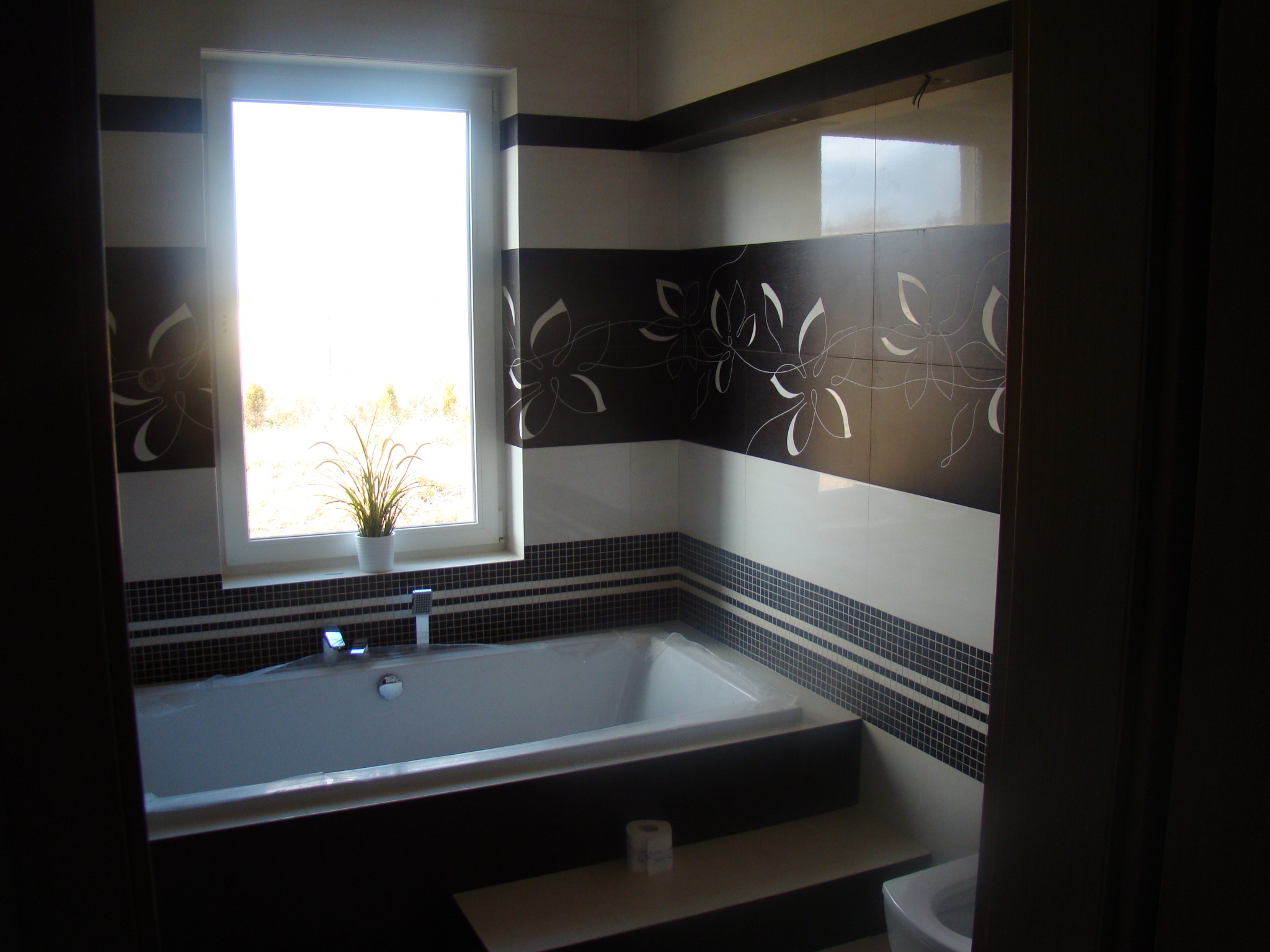 Nowoczesna łazienka z wanną pod oknem, wykończona płytkami w odcieniach bieli i brązu z motywem kwiatowym, widoczna toaleta i rolka papieru toaletowego na podeście.