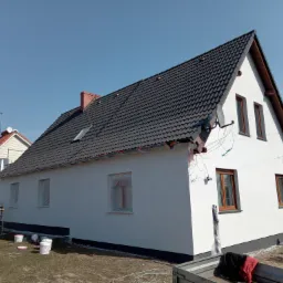 Dom jednorodzinny z nową, ciemną dachówką i świeżo pomalowaną elewacją na biało; okna zabezpieczone folią malarską, widoczne wiadra z farbą i narzędzia malarskie, fragment przyczepy; obok drugi dom...