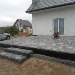 Wykończony taras z szarej kostki brukowej, z trzema stopniami wejściowymi, obok biały dom z szarym dachem i samochód osobowy w tle.