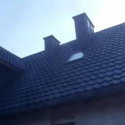 Dom z ciemną blachodachówką, trzema kominami i oknem dachowym, widok z dołu na tle pochmurnego nieba.