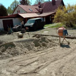 Przygotowanie podłoża pod przyszłą nawierzchnię z widoczną warstwą piasku, pomarańczową betoniarką i zaparkowanym busem firmy budowlanej na tle domu z czerwoną dach&oacute;wką.