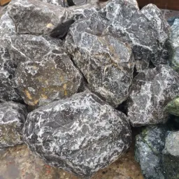 Kupa mokrego, czarno-białego węgla kamiennego o nieregularnych kształtach, z widocznymi smugami i przebarwieniami, leżąca na lekko wilgotnym podłożu.