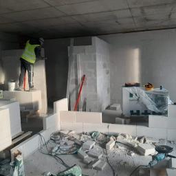 El-Sebo - Wnętrze w trakcie budowy ścian z białych bloczków, pracownik w kamizelce odblaskowej wyrównuje powierzchnię, widoczne narzędzia, worki po zaprawie oraz paczki bloczków betonowych.