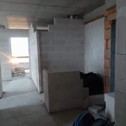 El-Sebo - Wnętrze w trakcie budowy: surowy betonowy strop, ściany z białych bloczków, fragment ściany z cegły, narzędzia budowlane w tle, wpadające światło przez okno.