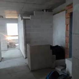 Wnętrze w trakcie budowy: surowy betonowy strop, ściany z białych bloczków, fragment ściany z cegły, narzędzia budowlane w tle, wpadające światło przez okno.