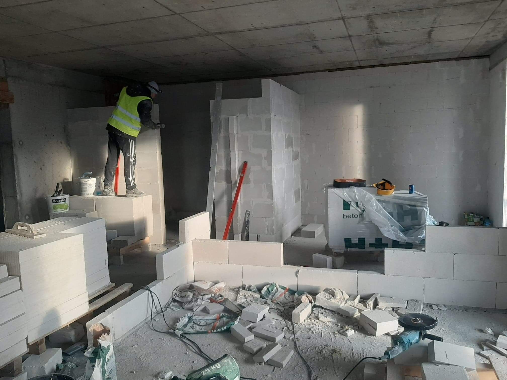 Wnętrze w trakcie budowy ścian z białych bloczków, pracownik w kamizelce odblaskowej wyrównuje powierzchnię, widoczne narzędzia, worki po zaprawie oraz paczki bloczków betonowych.