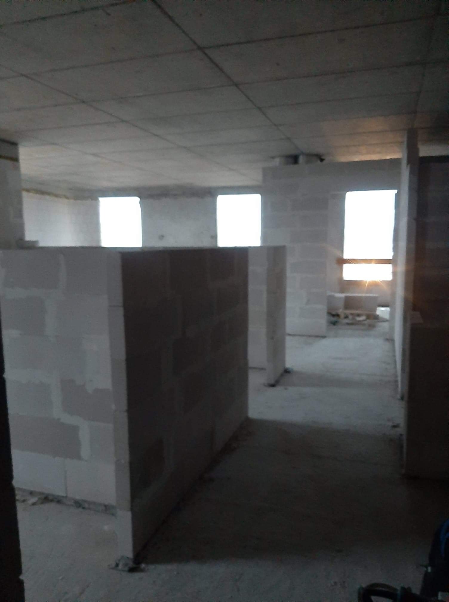 Wnętrze budynku w trakcie budowy z widocznymi ścianami z bloczków betonowych, surowym betonowym sufitem i oknami wpuszczającymi naturalne światło.