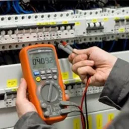 Elektryk mierzy napięcie multimetrem na tablicy rozdzielczej z bezpiecznikami, widoczne przewody i elementy instalacji elektrycznej.