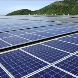 Rozległa instalacja paneli słonecznych na dachu budynku, z zielonym wzgórzem w tle pod błękitnym niebem.