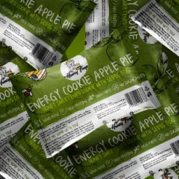 Zbliżenie na zielone opakowania ciastek energetycznych Honey Trap z jabłkiem i nasionami, ułożone warstwami, z widocznymi informacjami o składzie i wartościach odżywczych.