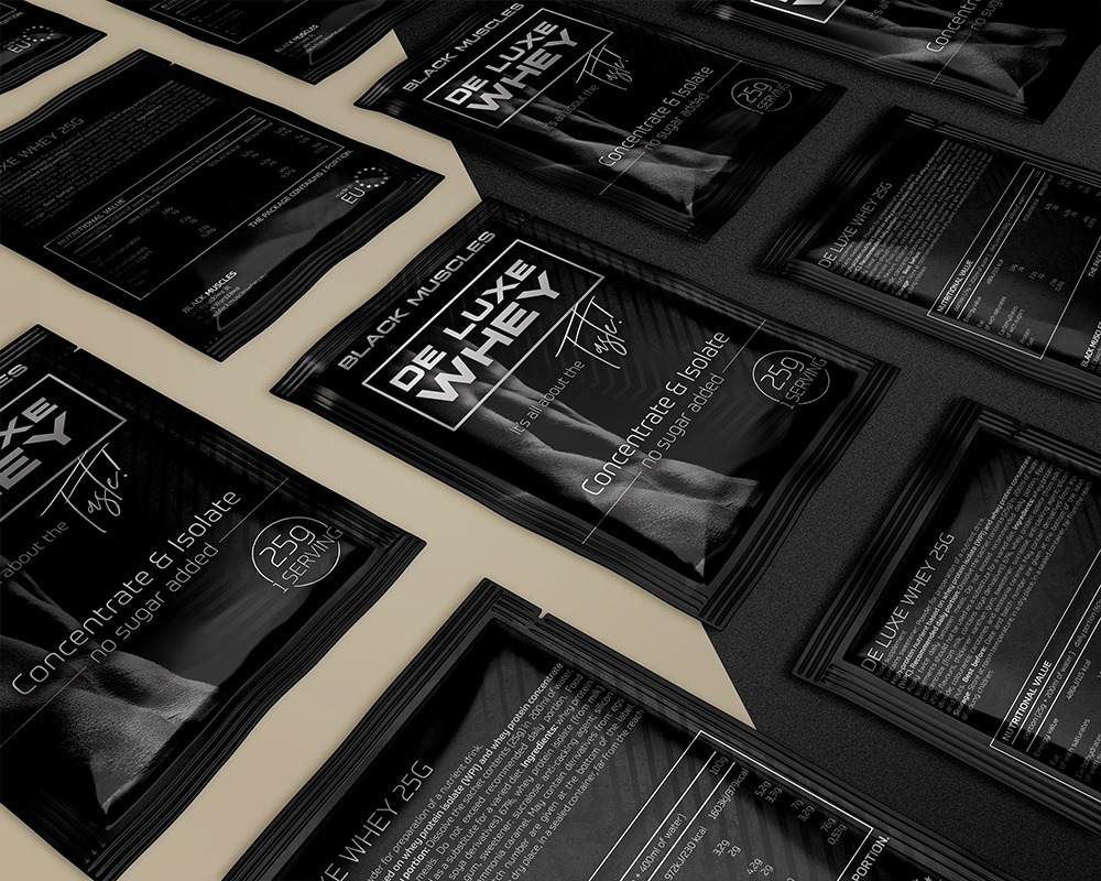 Rozłożone próbki czarnych, matowych saszetek z odżywką białkową Black Muscles De Luxe Whey, ułożone pod kątem na ciemnoszarym tle, eksponujące logo i informacje o wartościach odżywczych.