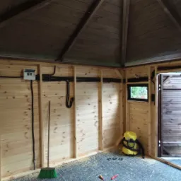 Wnętrze drewnianej altany z rozprowadzoną instalacją elektryczną: kable w peszlach, skrzynka rozdzielcza, małe okno, otwarte dwuskrzydłowe drzwi, w tle samochód.