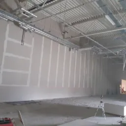 Montaż płyt karton-gips w przestronnym, komercyjnym wnętrzu z widocznymi instalacjami pod sufitem i laserowym poziomowaniem na ścianie.