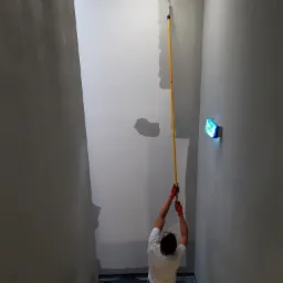 Pracownik w czerwonych rękawicach maluje ścianę wałkiem na długim kiju, widoczne ślady świeżej farby i zabezpieczona taśmą sufitowa linia.