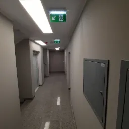 Długi korytarz w biurowcu z szarą podłogą z lastryko, beżowymi ścianami, oświetleniem LED i znakami ewakuacyjnymi.