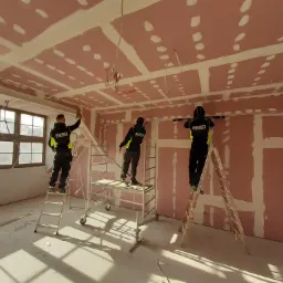 Trzech pracownik&oacute;w w firmowych uniformach na rusztowaniach i drabinie, wygładzających łączenia płyt kartonowo-gipsowych na suficie i ścianach w jasnym pomieszczeniu z dużymi oknami.
