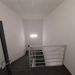 Wnętrze klatki schodowej z czarnymi płytkami na podłodze i białymi ścianami, widoczne ślady po przewodach na ścianie, oświetlenie sufitowe.