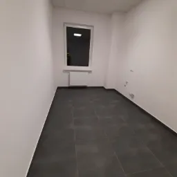 Puste, odświeżone wnętrze pokoju w Kwidzynie z ciemną podłogą z płytek, białymi ścianami i oknem z grzejnikiem. Widoczne gniazdka elektryczne.