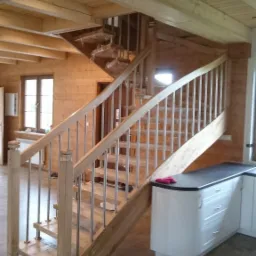 Jasne, drewniane schody z metalowymi balustradami w domu z drewnianymi ścianami i sufitem; spiralne schody w tle częściowo owinięte folią ochronną.