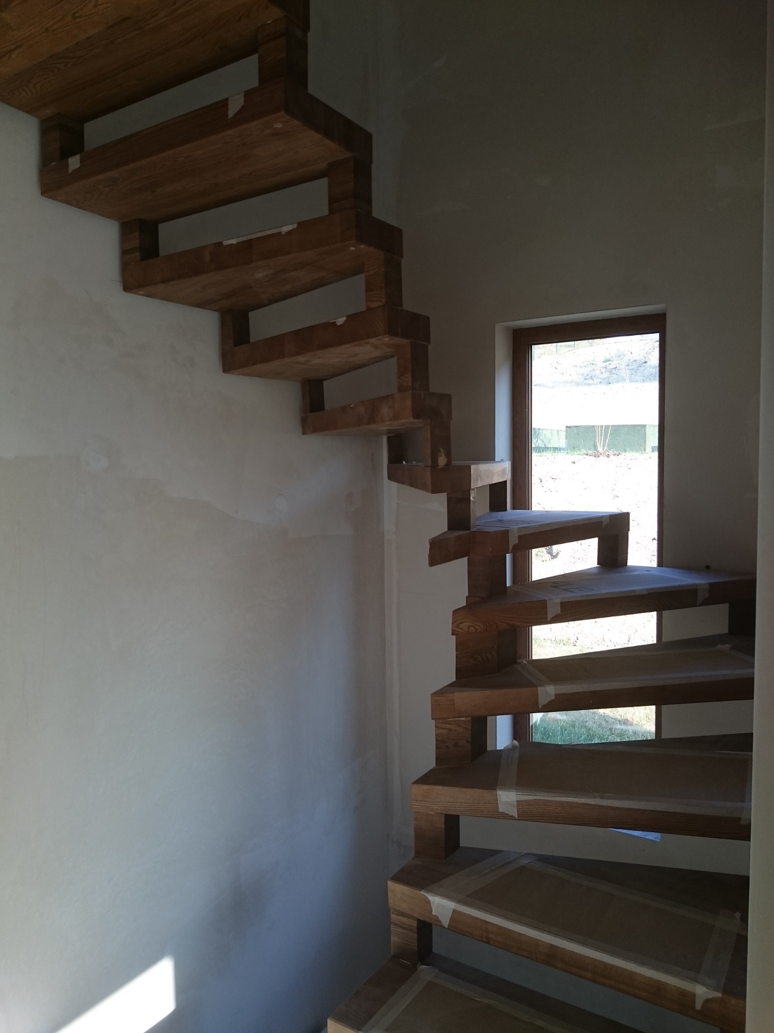 Kątowe, drewniane stopnie schodów bez podstopnic w surowym wnętrzu, zabezpieczone folią malarską, z widokiem na zewnątrz przez okno.