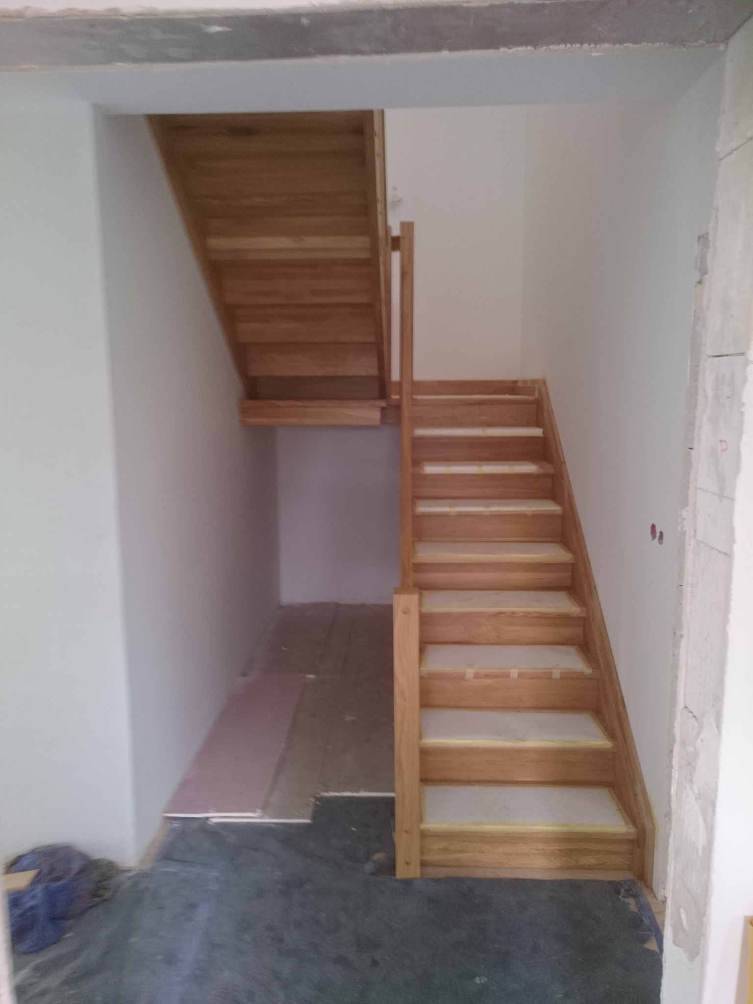 Drewniane schody z jasnego drewna podczas montażu, zabezpieczone taśmą malarską, widoczne surowe ściany i podłoga w trakcie wykończenia.
