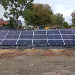 Rozległa instalacja paneli słonecznych w trakcie montażu na gruncie, widoczne czarne maty pod panelami, wystające słupki montażowe, rozsypane cegły, w tle domy i maszyny budowlane JCB.