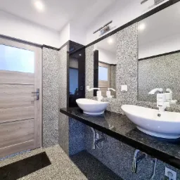 Łazienka z dwoma owalnymi umywalkami na czarnym blacie kamiennym, ściany wyłożone szarym kamieniem z widoczną teksturą, drewniane drzwi z przeszkleniem po lewej stronie.