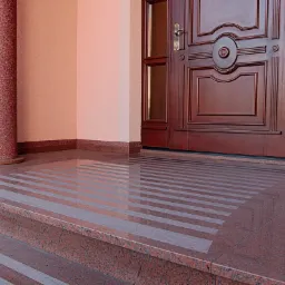 Polerowane, czerwono-szare, granitowe schody zewnętrzne z poziomymi, jaśniejszymi pasami, prowadzące do ciemnych, drewnianych drzwi wejściowych z kolumną po lewej stronie.