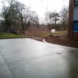 Świeżo wylany, gładki betonowy plac z widocznymi szczelinami dylatacyjnymi, obrzeżem z ziemi i fragmentami trawnika w tle, na terenie budowy w pochmurny dzień.