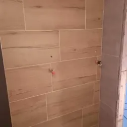 Ściana w trakcie układania płytek imitujących drewno, widoczne wystające rurki instalacji wodnej, szare płytki na krawędzi ściany.