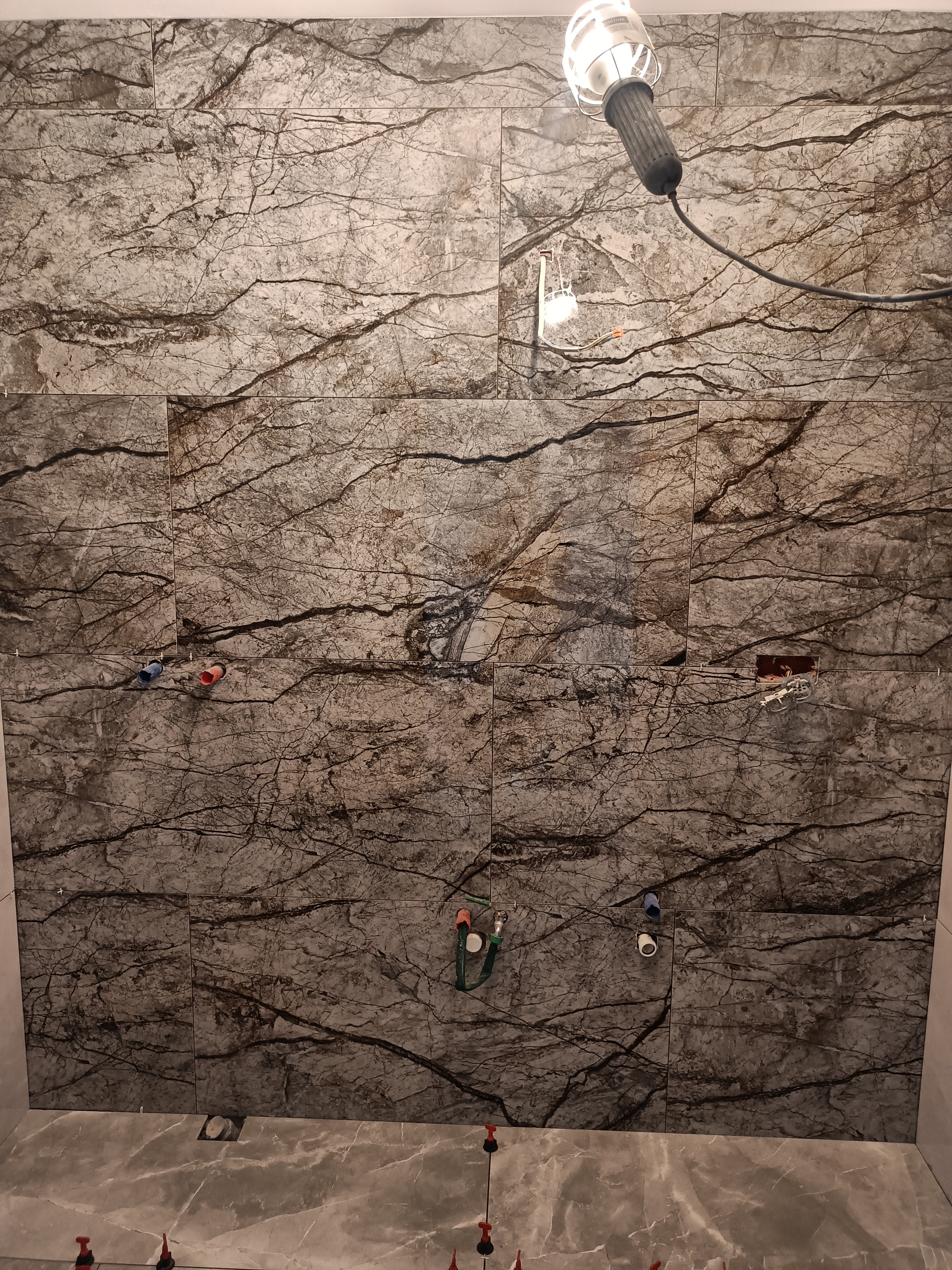 Montaż płytek imitujących marmur na ścianie w łazience, widoczne przyłącza wodne i lampa robocza.