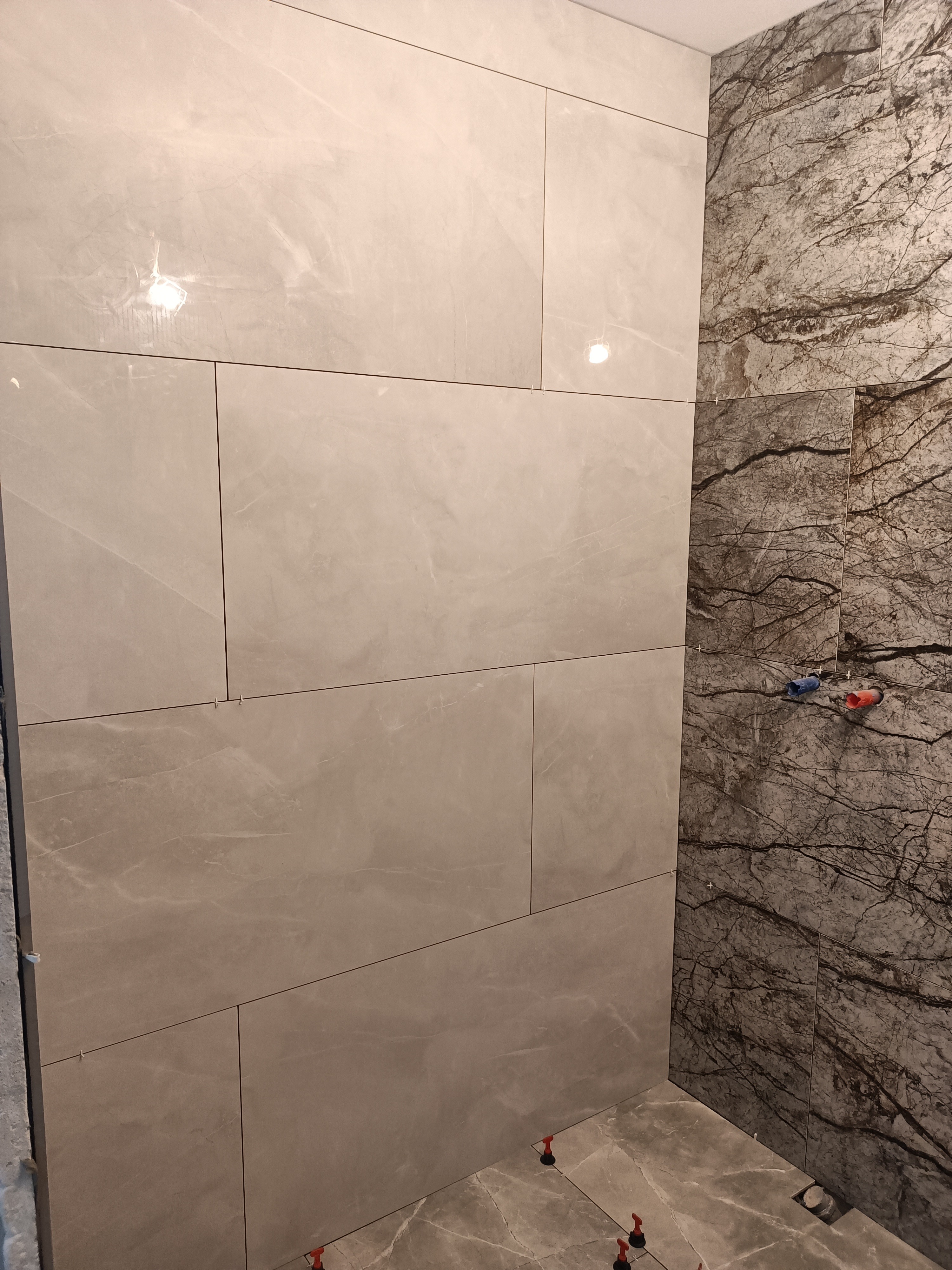 Łazienka w trakcie wykańczania: ściany wyłożone jasnymi, marmurkowymi płytkami wielkoformatowymi po lewej oraz ciemnymi płytkami imitującymi marmur z wyraźnym użyleniem po prawej stronie, widoczne...