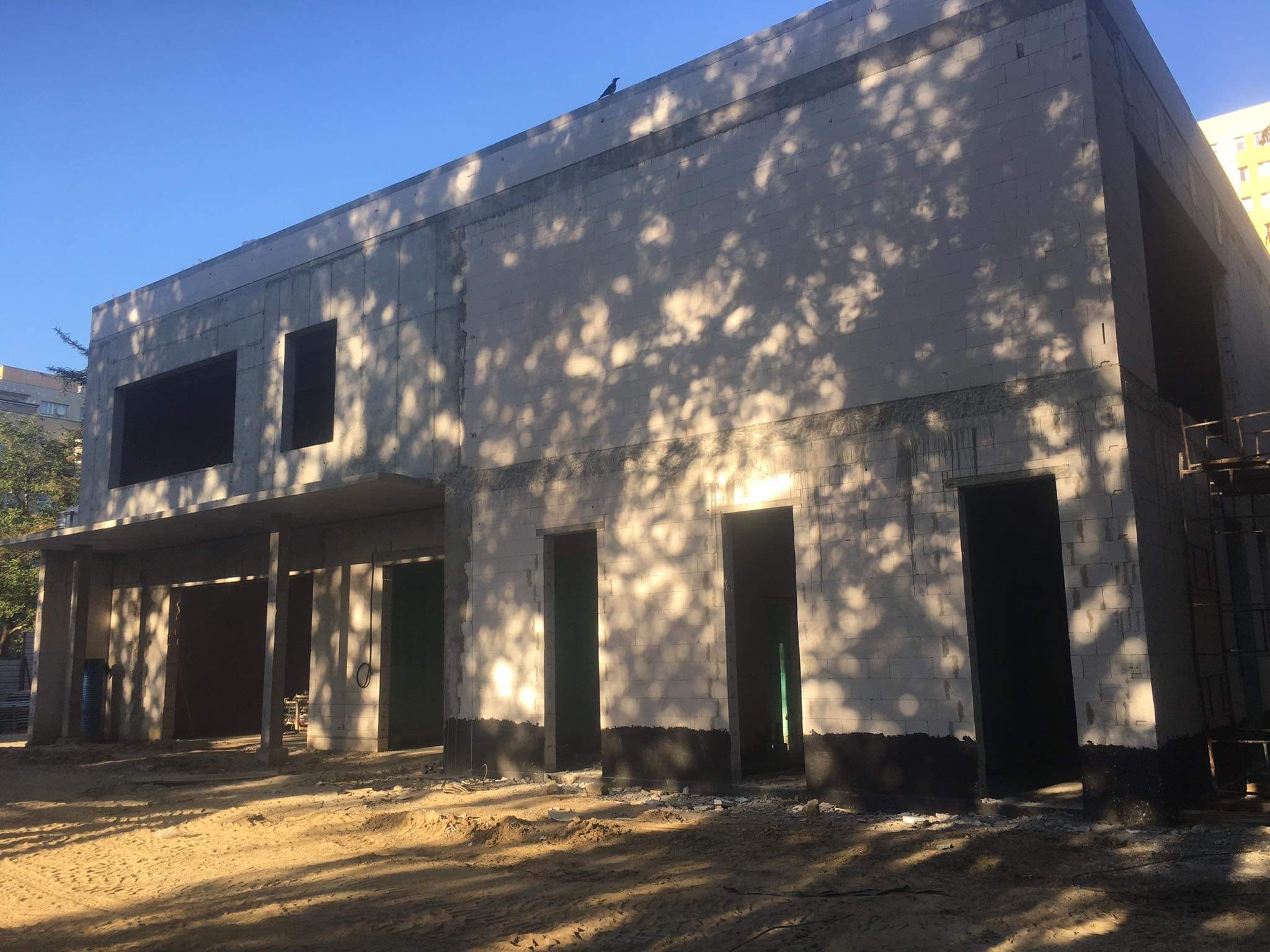 Surowy budynek w trakcie wznoszenia z białych bloczków, widoczne otwory okienne i drzwiowe, ziemia na zewnątrz, cień drzewa na ścianie, na dachu siedzi ptak.