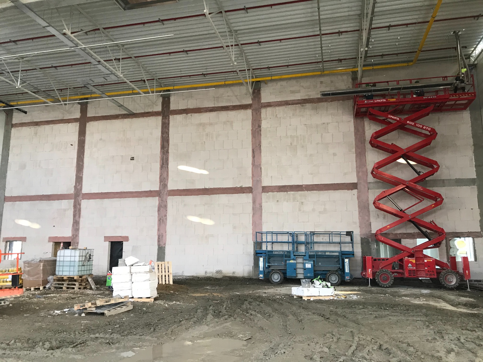 Wnętrze hali w stanie surowym z murem z bloczków i widocznymi śladami po szpachlowaniu na filarach, czerwony podnośnik nożycowy przy ścianie, niebieski podnośnik koszowy na dole, podłoże błotniste,...