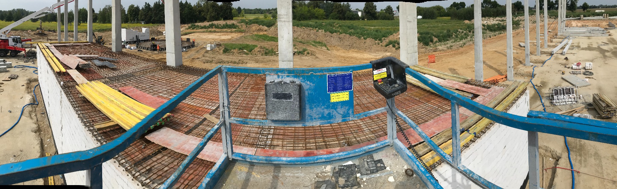 Widok z podnośnika na plac budowy z betonowymi słupami, zbrojeniami i deskowaniem, w tle krajobraz z zielenią.