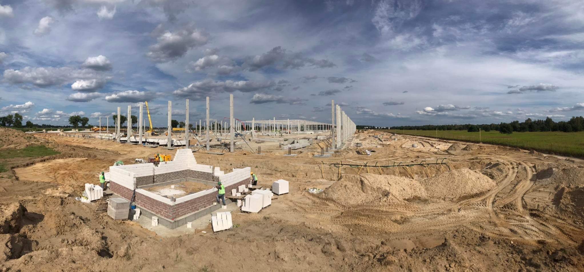 Szeroki kadr na plac budowy hali przemysłowej z widocznymi betonowymi słupami konstrukcyjnymi, fragmentem murowanej ściany z cegły silikatowej i pracownikami w kamizelkach odblaskowych.