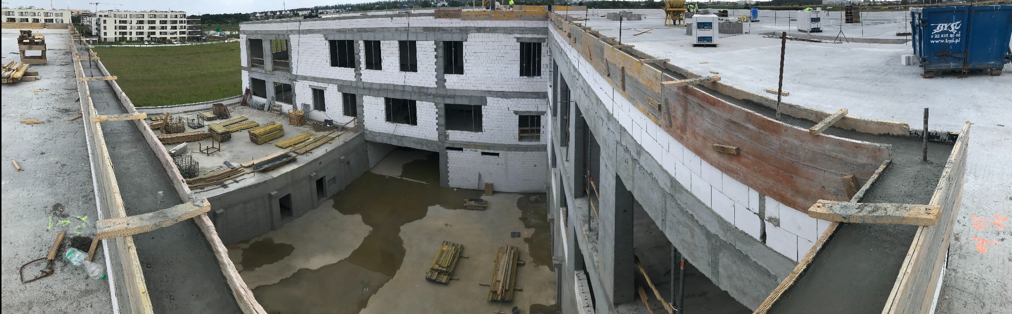 Panorama z góry przedstawia budynek w trakcie budowy z białego gazobetonu, widoczne szalunki zalane betonem, na dole zalana wodą piwnica, w tle budynki mieszkalne i zieleń.