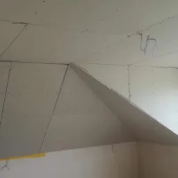 Montaż płyt gipsowo-kartonowych na poddaszu, widoczne łączenia płyt, wystające przewody elektryczne, okno dachowe w tle.