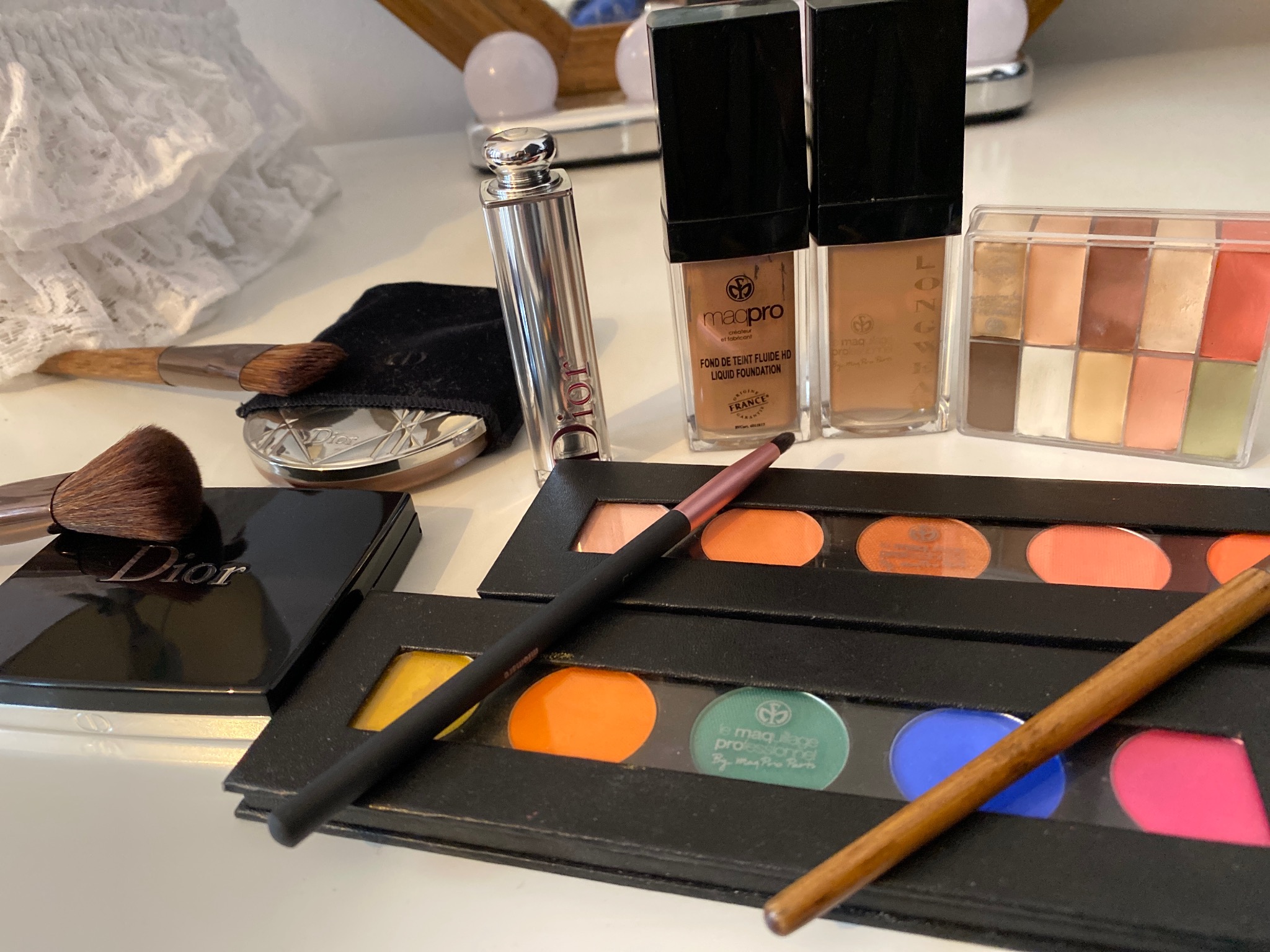 Zestaw kosmetyków do makijażu: palety cieni i róży, podkłady w butelkach, pędzle i puder marki Dior, ustawione na białym stole z lustrem z żarówkami w tle.