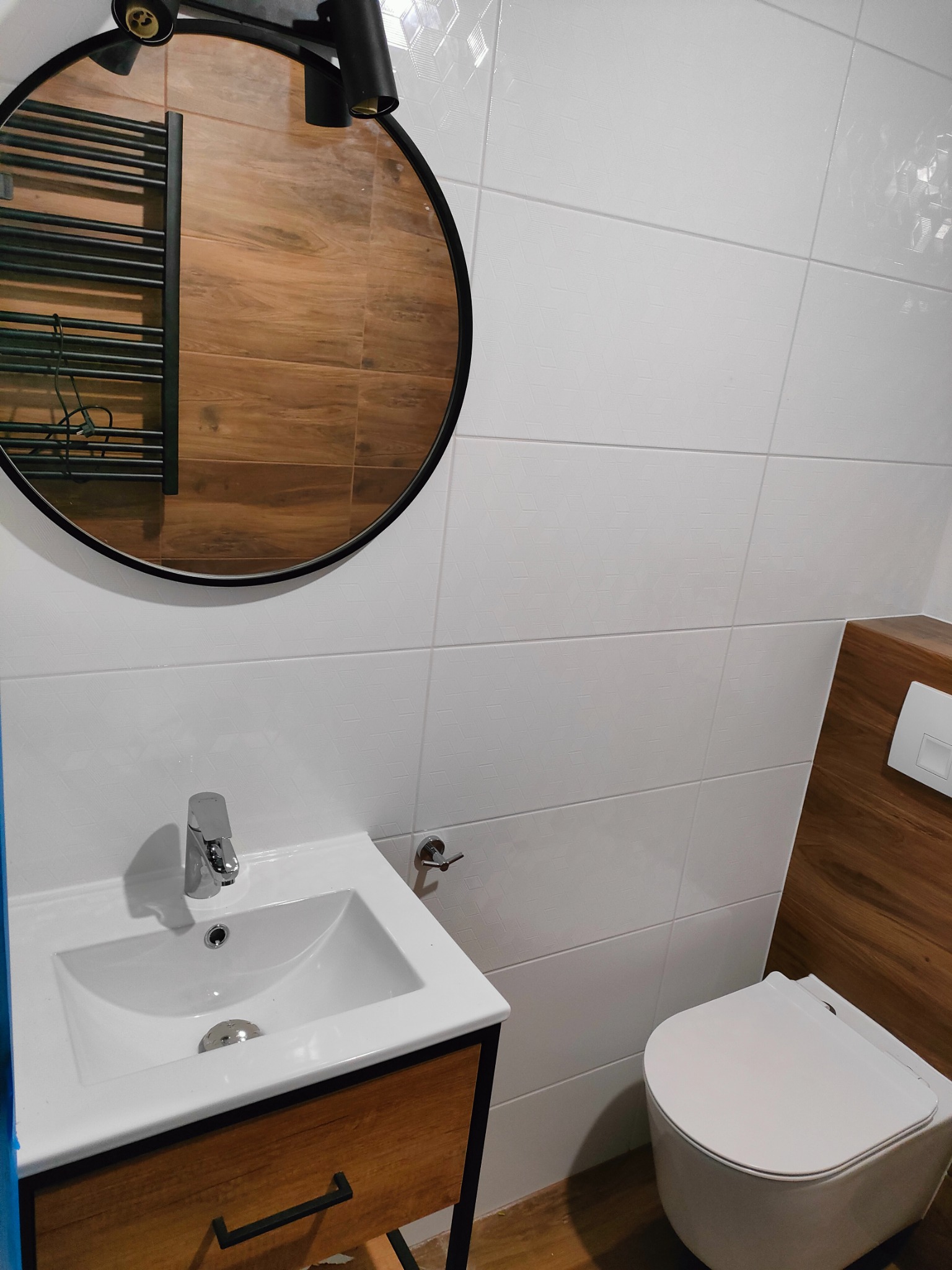 Nowoczesna łazienka z białą umywalką nablatową na drewnianej szafce z czarnymi uchwytami, okrągłe lustro z czarną ramą odbija grzejnik drabinkowy, toaleta w zabudowie z drewnianym akcentem, białe...