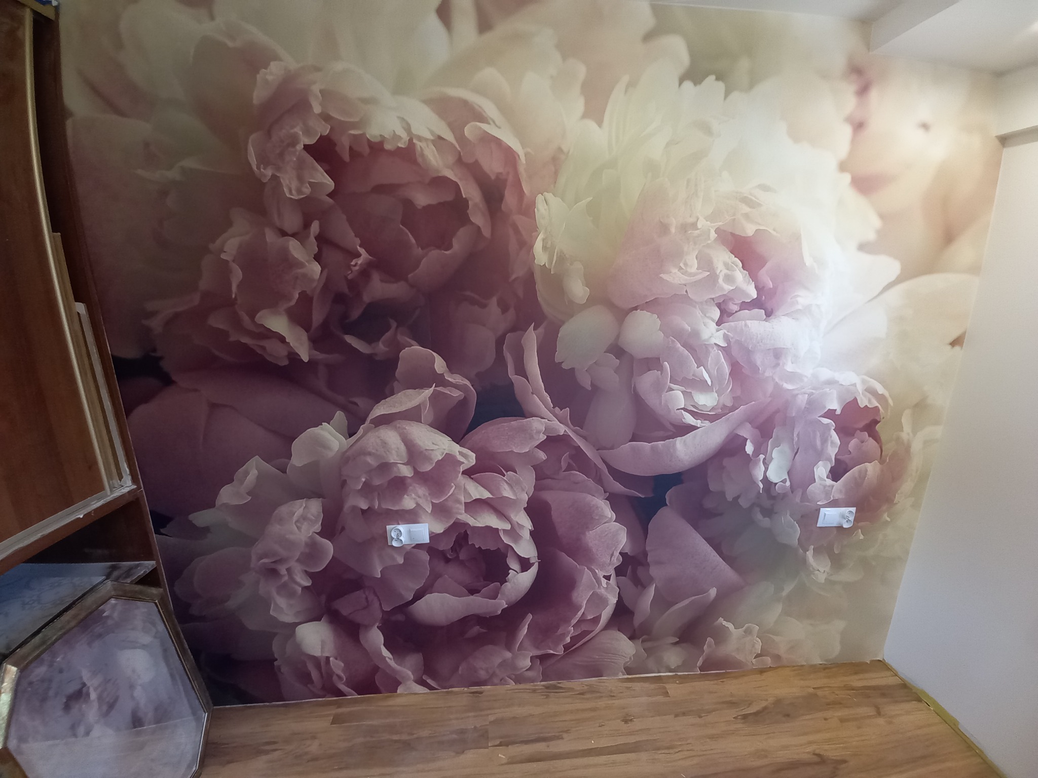 Fototapeta z motywem dużych, różowych piwonii na ścianie, widoczne gniazdka elektryczne i fragment drewnianej podłogi.