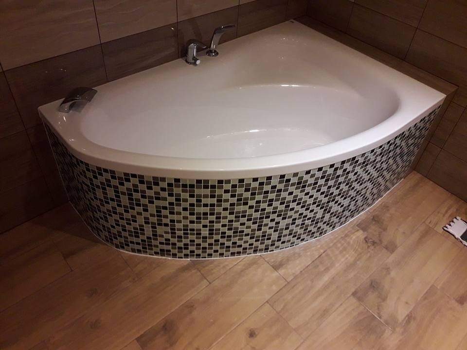 Narożna wanna obudowana mozaiką w odcieniach szarości i brązu, z drewnopodobną podłogą i elementami armatury.