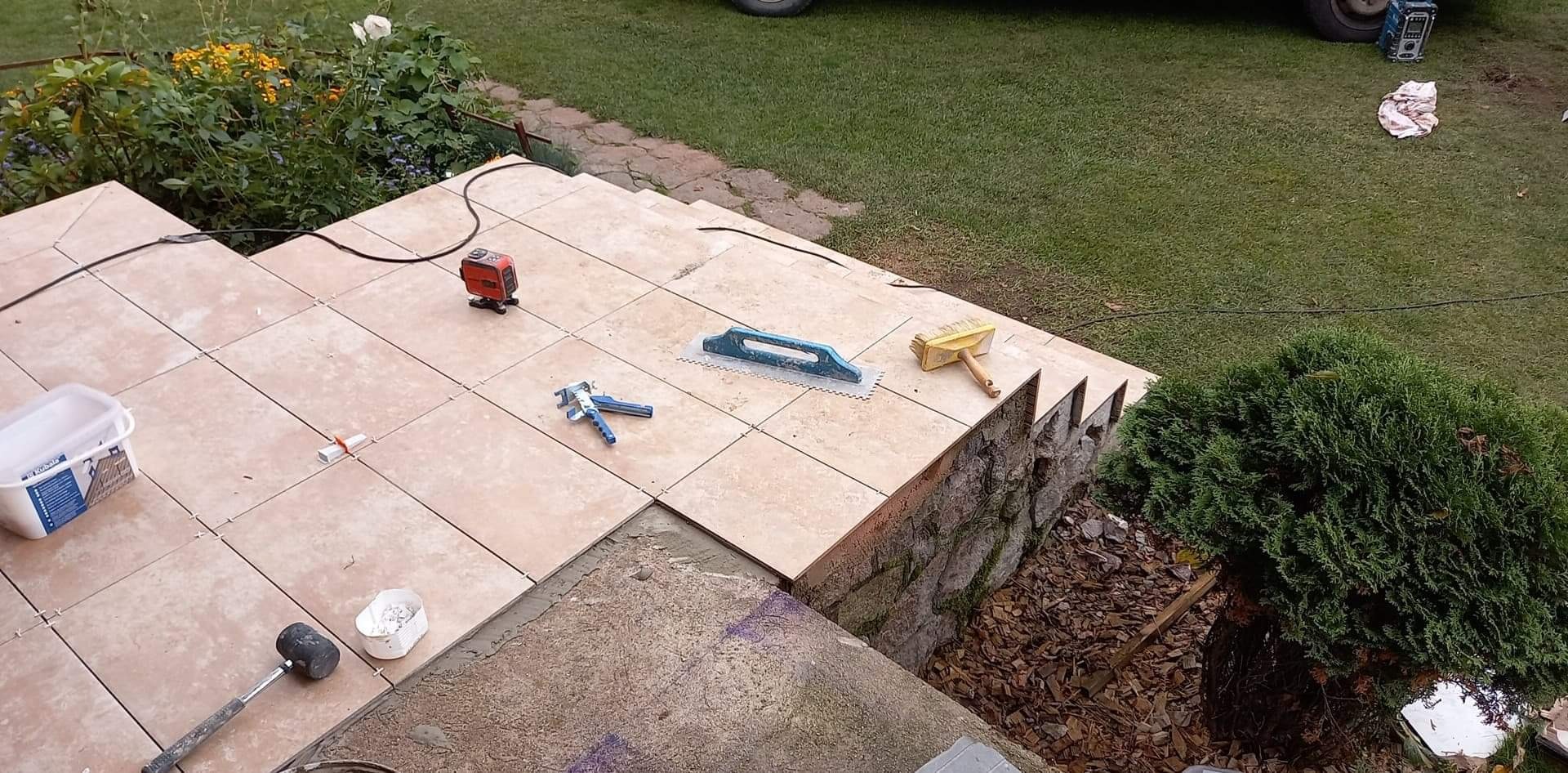 Układanie jasnych płytek na schodach zewnętrznych, narzędzia glazurnicze i poziomica laserowa na powierzchni, widoczna trawa w tle.